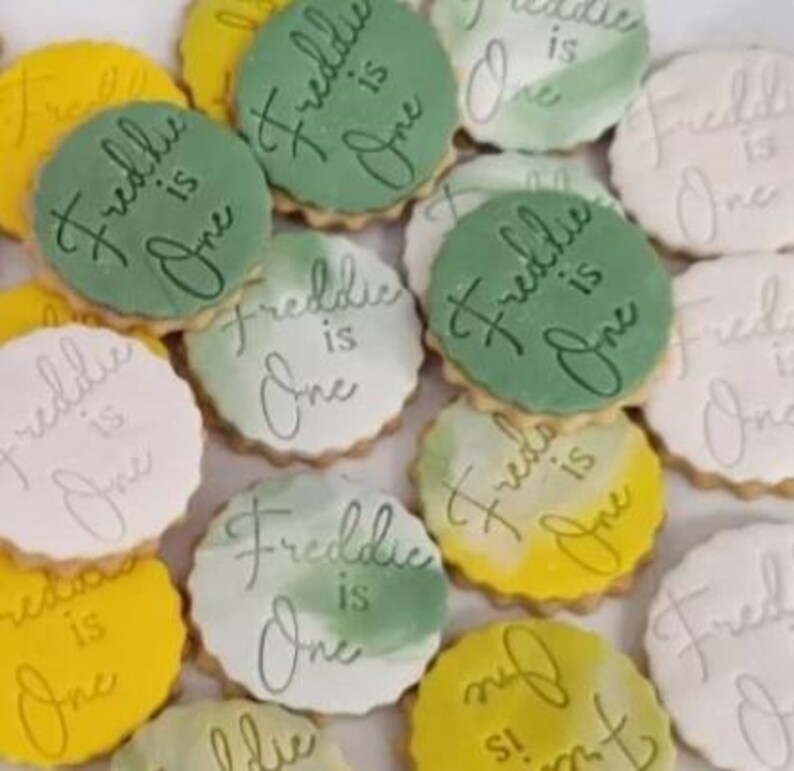 May include: A collection of decorated sugar cookies with green and yellow icing. The cookies are decorated with the text "Freddie is One" in white cursive writing.