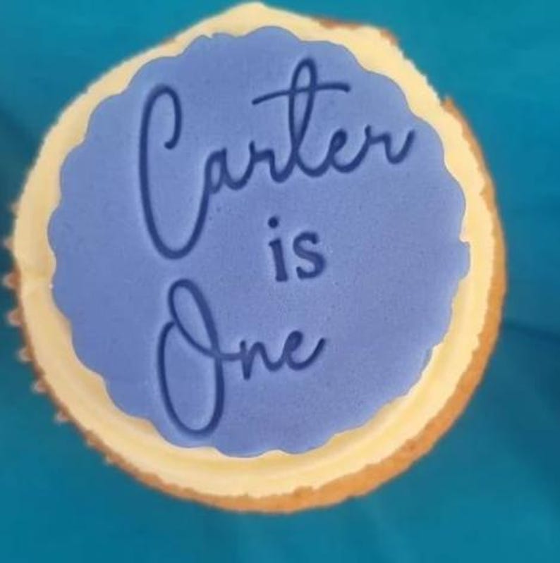 May include: A blue cupcake with white frosting and a blue fondant topper that says "Carter is One" in white script.