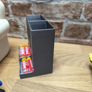 May include: A dark gray rectangular battery holder with two compartments. The holder contains several red and yellow Kodak AA batteries. The holder is on a wooden surface with a brick wall in the background.