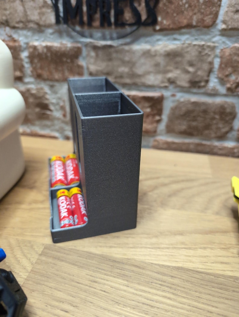 May include: A dark gray rectangular battery holder with two compartments. The holder contains several red and yellow Kodak AA batteries. The holder is on a wooden surface with a brick wall in the background.