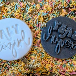 May include: Two circular objects with text and a bow design are displayed on a bed of colorful sprinkles. One is white and says "BABY girl". The other is dark gray and says "Yara Injo". Gold leaf accents are scattered around.