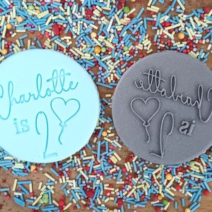May include: Two round, blue and gray silicone cake decorating stamps. The blue stamp says "Charlotte is 2" with a heart-shaped balloon. The gray stamp says "Charlotte is 2" with a heart-shaped balloon.