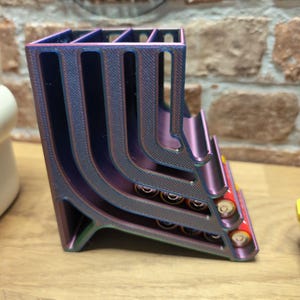 May include: A multi-colored, 3D-printed battery holder. The holder is a gradient of purple, blue, and green. It has multiple slots to hold batteries. The word "IMPRESS" is visible in the background.
