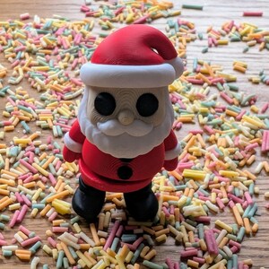 Op de afbeelding: Een rood en wit kerstmanfiguurtje dat op een houten oppervlak staat dat bedekt is met kleurrijke sprinkels. Het figuurtje heeft een witte baard en zwarte ogen.