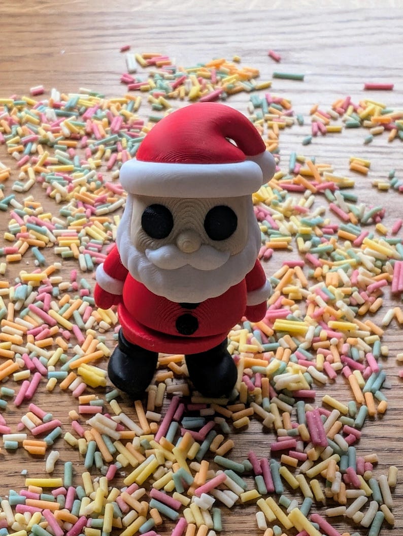 Op de afbeelding: Een rood en wit kerstmanfiguurtje dat op een houten oppervlak staat dat bedekt is met kleurrijke sprinkels. Het figuurtje heeft een witte baard en zwarte ogen.