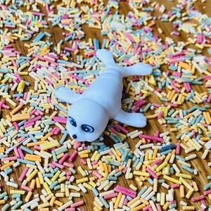 May include: A white ceramic seal figurine with blue eyes lying on a surface covered in colorful sprinkles.