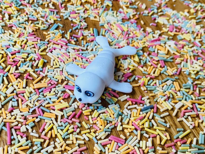 May include: A white ceramic seal figurine with blue eyes lying on a surface covered in colorful sprinkles.