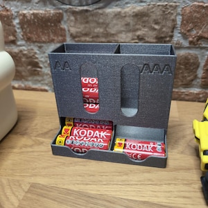 May include: A gray 3D-printed battery organizer with compartments for AA and AAA batteries. The organizer holds red and yellow Kodak batteries. The word "KODAK" is visible on the batteries.