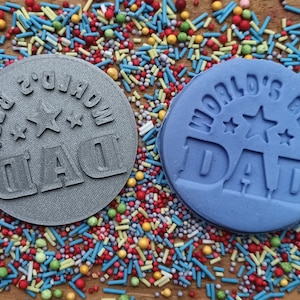 May include: Two round, embossed decorations with the text "WORLD'S BEST DAD" and star accents. One is gray, the other is blue. The decorations are surrounded by colorful sprinkles.