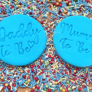 May include: Two blue round silicone molds with the text "Daddy to Be" and "Mummy to Be" in white script with a small heart symbol next to each phrase. The molds are on a surface covered in colorful sprinkles.