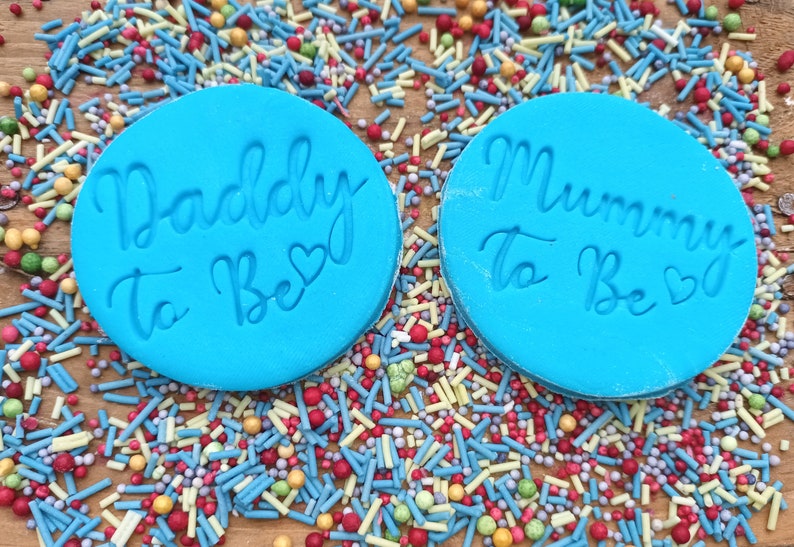 May include: Two blue round silicone molds with the text "Daddy to Be" and "Mummy to Be" in white script with a small heart symbol next to each phrase. The molds are on a surface covered in colorful sprinkles.