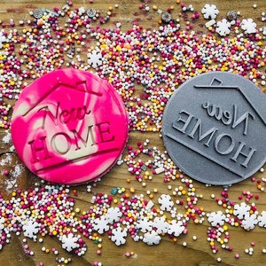May include: A pink and white swirled cookie with the words "New Home" and a house outline. A gray cookie stamp with the words "New Home" in reverse. The cookies are surrounded by colorful sprinkles and snowflake-shaped decorations.