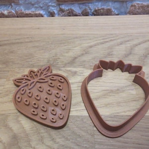 May include: Two brown strawberry-shaped cookie cutters. One cutter is a solid strawberry shape with detailed seed impressions. The other is a strawberry outline with a leaf-like top. Both are made of plastic and sit on a wooden surface.