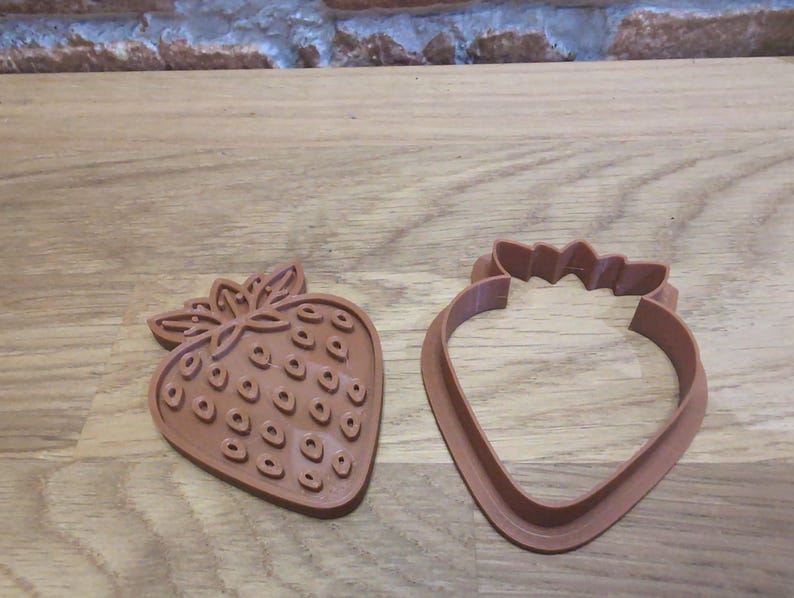 May include: Two brown strawberry-shaped cookie cutters. One cutter is a solid strawberry shape with detailed seed impressions. The other is a strawberry outline with a leaf-like top. Both are made of plastic and sit on a wooden surface.