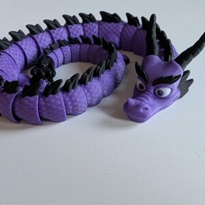 K&ouml;nnte beinhalten: Ein lila und schwarzes, segmentiertes Drachenspielzeug mit einer strukturierten Oberfl&auml;che. Der Drache hat einen gro&szlig;en Kopf mit schwarzen Augen und einen missmutigen Gesichtsausdruck.