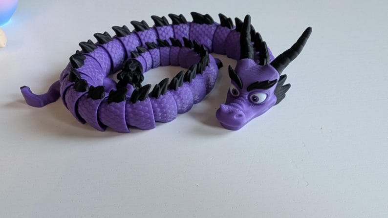K&ouml;nnte beinhalten: Ein lila und schwarzes, segmentiertes Drachenspielzeug mit einer strukturierten Oberfl&auml;che. Der Drache hat einen gro&szlig;en Kopf mit schwarzen Augen und einen missmutigen Gesichtsausdruck.