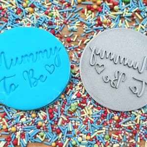 May include: Two round blue and gray silicone stamps with the text "Mummy to Be" and "Daddy to Be" in white and gray lettering. The stamps are surrounded by colorful sprinkles.