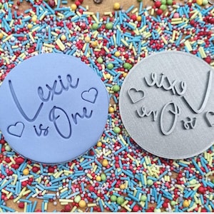 May include: Two round, blue and gray cake decorating stamps with the text "Lexie is One" in a cursive font. The stamps are on a background of colorful sprinkles.