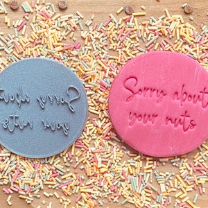 May include: Two round, gray and pink cookie cutters with the text "Sorry about your mug" and "Sorry about your nuts" on them. The cookie cutters are on a wooden surface with colorful sprinkles.