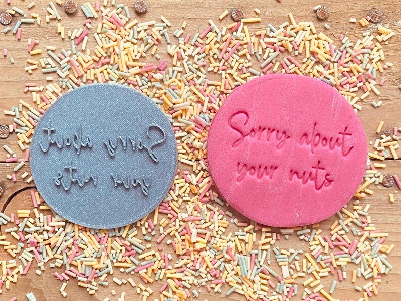 May include: Two round, gray and pink cookie cutters with the text "Sorry about your mug" and "Sorry about your nuts" on them. The cookie cutters are on a wooden surface with colorful sprinkles.