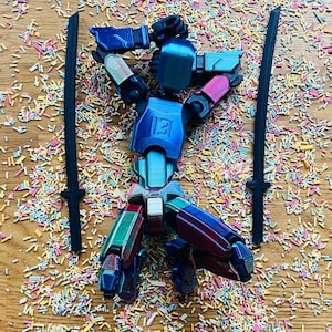 May include: A multi-colored robot toy with two black swords, set against a background of colorful sprinkles. The robot has blue, red, green, and white accents. The letter "E" is visible on the robot's chest.