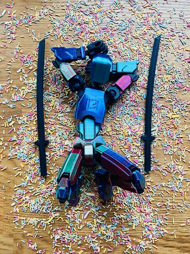 May include: A multi-colored robot toy with two black swords, set against a background of colorful sprinkles. The robot has blue, red, green, and white accents. The letter "E" is visible on the robot's chest.