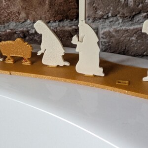 May include: A gold and cream-colored nativity scene. The figures are cut-out silhouettes, including Mary, Joseph, the baby Jesus in a manger, and the three wise men. The figures are mounted on a gold-colored base.