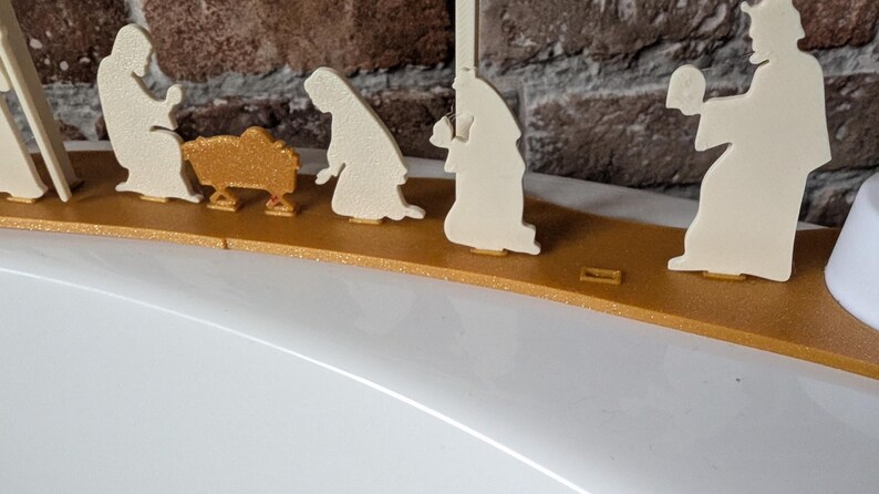 May include: A gold and cream-colored nativity scene. The figures are cut-out silhouettes, including Mary, Joseph, the baby Jesus in a manger, and the three wise men. The figures are mounted on a gold-colored base.