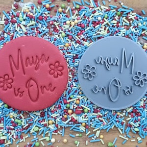 May include: Two round, red and gray, silicone cake decorating stamps with the text "Maya is One" in a cursive font. The stamps are surrounded by colorful sprinkles.