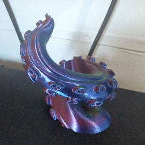 May include: A 3D-printed sculpture of an octopus tentacle in shades of blue, purple, and red. The tentacle curves upwards, featuring textured suckers and a detailed, organic design. The sculpture is on a dark surface.