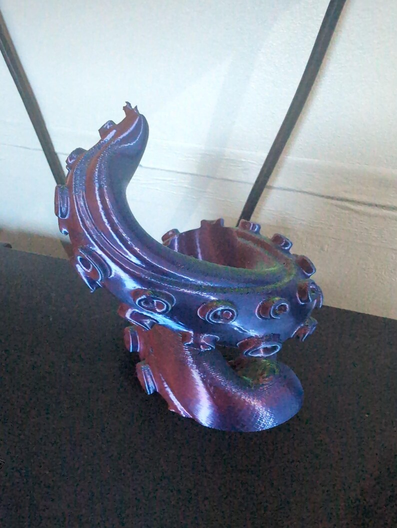 May include: A 3D-printed sculpture of an octopus tentacle in shades of blue, purple, and red. The tentacle curves upwards, featuring textured suckers and a detailed, organic design. The sculpture is on a dark surface.
