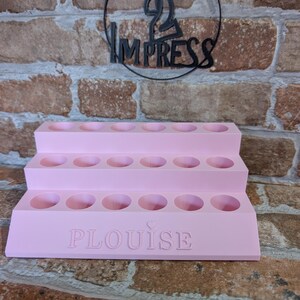 May include: A tiered, light pink display stand with multiple circular openings. The word "PLouise" is embossed on the front. Above the stand, a black metal sign reads "PRINT 2 IMPRESS" in a circular design. The background is a brick wall.