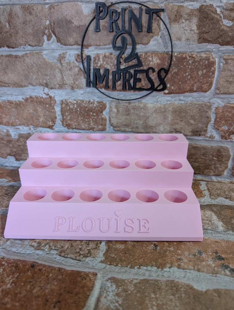 May include: A tiered, light pink display stand with multiple circular openings. The word "PLouise" is embossed on the front. Above the stand, a black metal sign reads "PRINT 2 IMPRESS" in a circular design. The background is a brick wall.