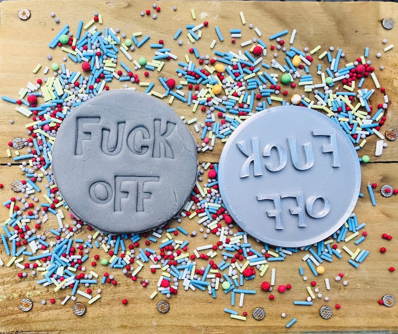 Op de afbeelding: Twee grijze, ronde koekjesstempels met de woorden "FUCK OFF" erop gedrukt. E&eacute;n stempel heeft de tekst in de juiste richting, de andere is omgekeerd. De stempels zijn omgeven door kleurrijke hagelslag op een houten oppervlak.