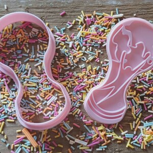 Może przedstawiać: Dwa r&oacute;żowe plastikowe wykrawacze do ciastek w kształcie butelki dla dziecka i twarzy dziecka, otoczone kolorową posypką na drewnianej powierzchni. Wykrawacze przeznaczone są do pieczenia i dekorowania ciasteczek.