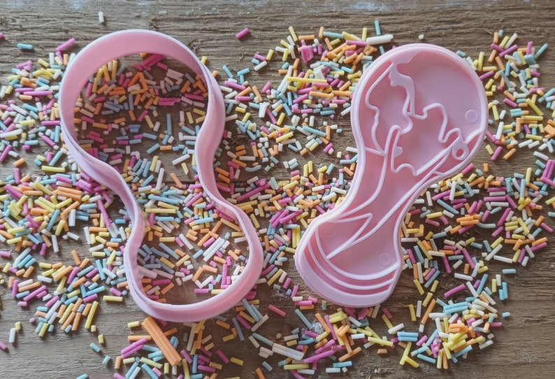 Może przedstawiać: Dwa r&oacute;żowe plastikowe wykrawacze do ciastek w kształcie butelki dla dziecka i twarzy dziecka, otoczone kolorową posypką na drewnianej powierzchni. Wykrawacze przeznaczone są do pieczenia i dekorowania ciasteczek.
