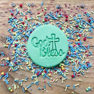 May include: A round, light green cookie with the words "God Bless" and a cross design pressed into the dough. The cookie is surrounded by colorful sprinkles on a wooden surface.