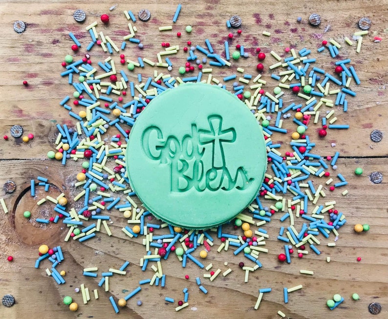 May include: A round, light green cookie with the words "God Bless" and a cross design pressed into the dough. The cookie is surrounded by colorful sprinkles on a wooden surface.