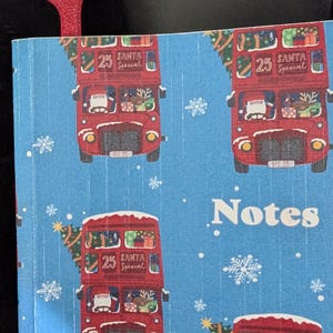 Pode incluir: Um caderno azul com um padr&atilde;o repetido de &ocirc;nibus vermelhos de dois andares decorados para o Natal. Os &ocirc;nibus t&ecirc;m o texto "25 Santa Special". Uma marca de p&aacute;gina vermelha em forma de m&atilde;o &eacute; vis&iacute;vel.