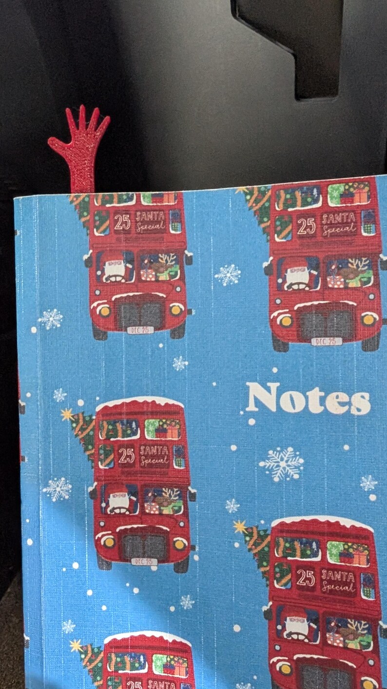 Pode incluir: Um caderno azul com um padr&atilde;o repetido de &ocirc;nibus vermelhos de dois andares decorados para o Natal. Os &ocirc;nibus t&ecirc;m o texto "25 Santa Special". Uma marca de p&aacute;gina vermelha em forma de m&atilde;o &eacute; vis&iacute;vel.