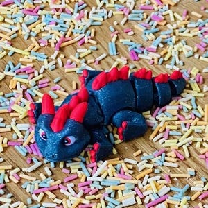 May include: A black and red fondant dragon figurine with a sparkly finish, sitting on a surface covered in colorful sprinkles.