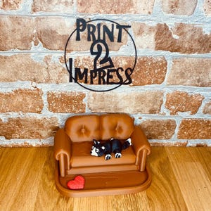 May include: A miniature brown sofa with a black and white dog figurine and a red heart. The sofa has a button-tufted design. The background features a brick wall and a black metal sign that reads "PRINT 2 IMPRESS".