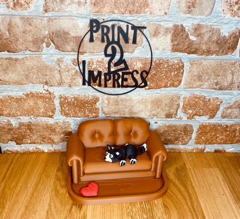 May include: A miniature brown sofa with a black and white dog figurine and a red heart. The sofa has a button-tufted design. The background features a brick wall and a black metal sign that reads "PRINT 2 IMPRESS".
