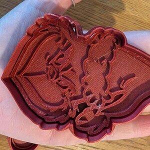 K&ouml;nnte beinhalten: Ein roter Ausstecher aus Kunststoff in Form einer Silhouette eines Paares. Der Ausstecher wird in einer Hand gehalten und das Design ist detailliert mit floralen Elementen. Der Ausstecher besteht aus rotbraunem Kunststoff.