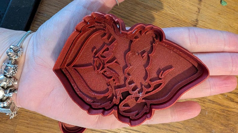 K&ouml;nnte beinhalten: Ein roter Ausstecher aus Kunststoff in Form einer Silhouette eines Paares. Der Ausstecher wird in einer Hand gehalten und das Design ist detailliert mit floralen Elementen. Der Ausstecher besteht aus rotbraunem Kunststoff.