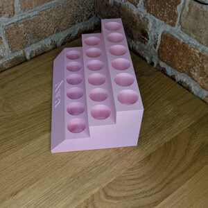 May include: A light pink tiered organizer with multiple circular holes. The organizer has the name "Ava" printed on the side. The organizer is made of a solid material and is designed to hold small items.