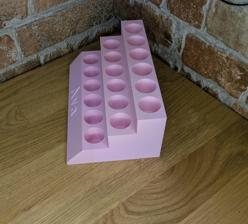 May include: A light pink tiered organizer with multiple circular holes. The organizer has the name "Ava" printed on the side. The organizer is made of a solid material and is designed to hold small items.