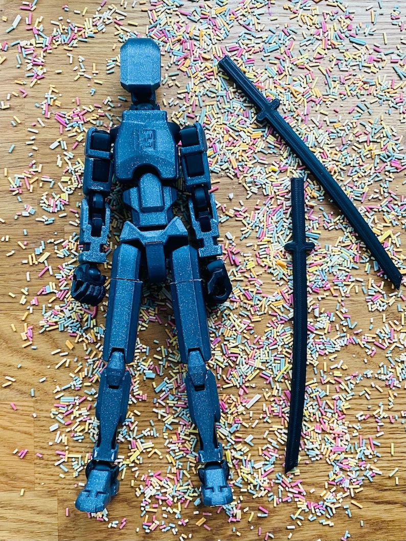 May include: A dark blue articulated action figure with a matching sword and a second sword piece. The figure is posed on a wooden surface covered in colorful sprinkles. The figure is approximately 6 inches tall.