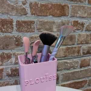 May include: A pink makeup brush holder with the name "PLouise" embossed on the front. The holder contains several makeup brushes with black, pink, and white bristles. The background is a brick wall.