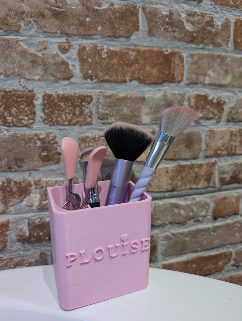 May include: A pink makeup brush holder with the name "PLouise" embossed on the front. The holder contains several makeup brushes with black, pink, and white bristles. The background is a brick wall.